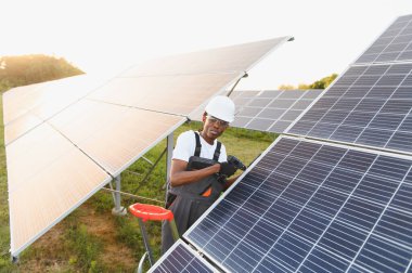 Güneş panelleri üzerinde çalışan genç siyah teknisyen, bir güneş parkına bir elektrikli alet kullanarak yenilenebilir enerji sistemi kuruyor.