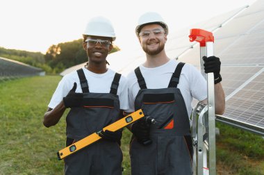 Gülümseyen çeşitli teknisyenler güneş panelleri yakınında araçlarla poz vererek yeşil enerjiyi, takım çalışmasını ve sürdürülebilir gücü sembolize ediyorlar.
