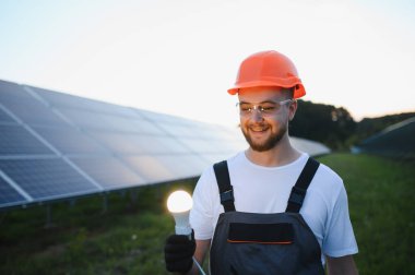 Şapkalı ve iş kıyafetli genç adam elinde parlayan bir ampul tutuyor, yenilenebilir enerjiyi ve parlak fikirleri simgeliyor.