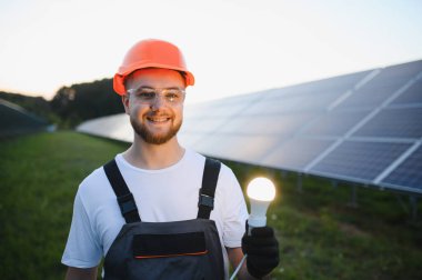 Gülümseyen teknisyen elinde parlak bir ampul tutuyor, temiz enerjiyi ve güneş enerjisi santralinde sürdürülebilir yeniliği simgeliyor.