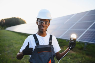 Gülümseyen teknisyen yenilenebilir enerji sergiliyor, arka planda çalışan güneş panelleri olan parlayan bir ampul tutuyor.