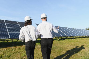 Sert şapkalı mühendisler bir alandaki güneş paneli dizisini inceleyerek yenilenebilir enerji ve sürdürülebilir enerjiyi tartışıyorlar.