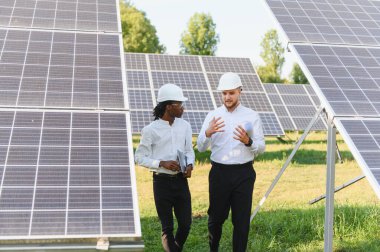 Güneş enerjisi santralinde yenilenebilir enerji projesini tartışan iki farklı kasklı ve gömlekli mühendis.