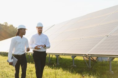 İki farklı mühendis, ellerinde planlar ve bir dizüstü bilgisayar, bir enerji santralinde yürüyorlar ve yenilenebilir güneş enerjisini tartışıyorlar.