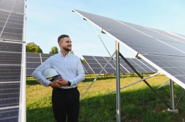 Güneş paneli alanında duran mühendis, elinde bir şapka tutuyor ve sürdürülebilir enerji kurulumunu gözlemliyor.