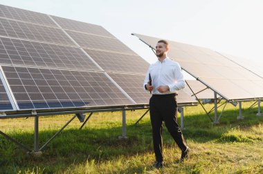 Elinde tablet tutan genç adam, yenilenebilir enerjiyi ve sürdürülebilir geleceği sembolize eden büyük bir güneş panelinin yanında güvenle yürüyor.