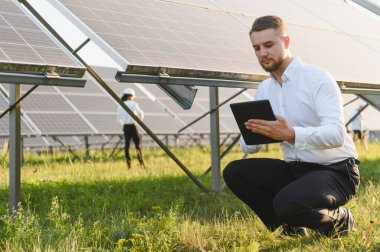 Erkek mühendis dijital tablet kullanarak güneş paneli sistemini inceliyor ve arka planda sürdürülebilir teknolojiyi sembolize eden bir meslektaşı var.