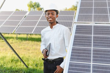 Elinde tablet olan Afrikalı Amerikalı adam güneş panellerinin arasında duruyor ve yenilenebilir enerji geliştirme üzerinde çalışıyor.