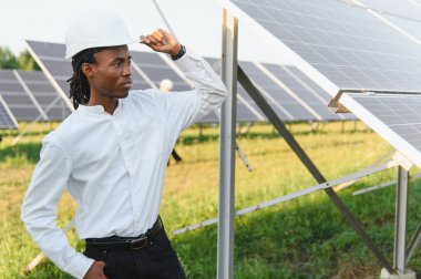 Sert bir şapka takan genç adam yenilenebilir enerji çiftliğindeki güneş panellerini kontrol ediyor ve sürdürülebilirliğe odaklanıyor.