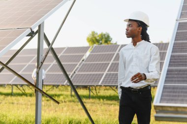 Gündüz vakti yenilenebilir bir enerji çiftliğinde şapka takan, yürüyen ve güneş panellerini inceleyen genç bir mühendis.