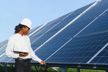 Siyah şapkalı ve elinde tablet olan Afrikalı Amerikalı adam, yenilenebilir enerji için büyük güneş panellerini kontrol ediyor.