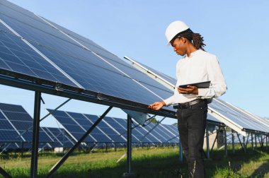 Siyah şapkalı Afrikalı erkek mühendis fotovoltaik bir çiftlikteki büyük güneş panellerini inceleyerek dijital tablet tutuyor.