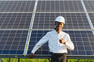 Sert bir şapka takan, gülümseyen ve baş parmak işareti yapan genç bir adam, güneş panellerinin önünde duruyor.