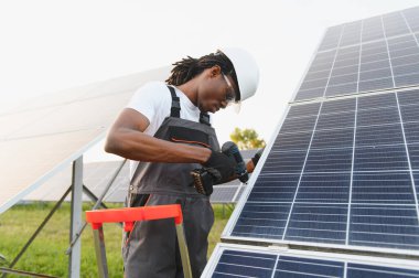 Siyah şapka ve güvenlik gözlüğü takan Afrikalı Amerikalı işçiler güneş panelini güneş çiftliğinde elektrikli aletlerle kuruyorlar.