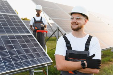 Kollarını kavuşturmuş gülümseyen mühendis, arka planda çalışan meslektaşım. Güneş çiftliği ekipmanlarını kontrol eden profesyoneller