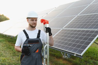 Beyaz kasklı ve tulumlu bir erkek işçi elinde matkap, bir elektrik santraline güneş panelleri kuruyor.