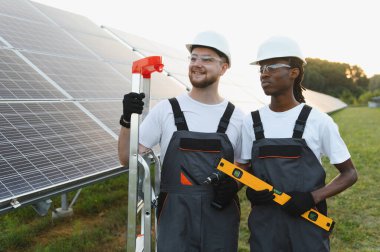 Bir güneş çiftliğinde duran iki farklı mühendis, ellerinde yenilenebilir enerji sistemi kurulumu ve bakımına hazır araçlar tutuyorlar.