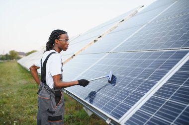 Genç siyahi teknisyen güneş panellerini cam sileceğiyle temizliyor, sürdürülebilir enerji üretimi için bakım yapıyor.