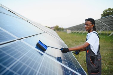 Rastalı bir adam güneş panelini uzun bir fırçayla temizliyor, büyük bir güneş enerjisi santralinde bakım yapıyor.