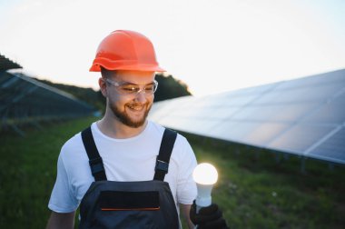 Sert şapkalı ve güvenlik gözlüklü genç bir adam güneş çiftliğinde parlayan bir ampul tutuyor, yenilenebilir enerji fikirlerini temsil ediyor.