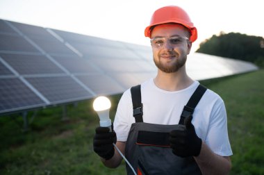 Kasketli ve koruyucu gözlüklü genç bir adam arka planda güneş panelleri olan parlayan bir ampul tutuyor.