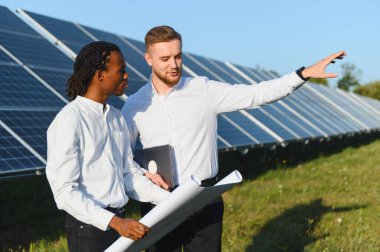 Bir güneş çiftliğinde iş birliği yapan mühendisler, planları gözden geçirir ve geniş bir açık havada yenilenebilir enerji çözümlerini tartışırlar.