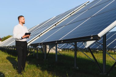 Bir güneş enerjisi santralini denetleyen erkek mühendis yenilenebilir enerji için dijital tabletle elektrik üretimini kontrol ediyor.