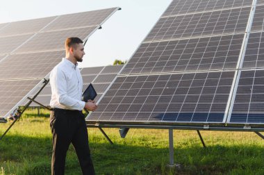 Bir güneş enerjisi çiftliğindeki sıra sıra fotovoltaik panelleri elinde tutan, yürüten ve denetleyen genç bir mühendis.