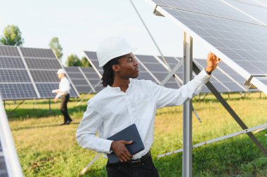 Sert şapka takan Afrikalı erkek mühendis güneş fotovoltaik panellerini kontrol ederek bir alanda yenilenebilir enerji sistemi çalışmasını sağlıyor