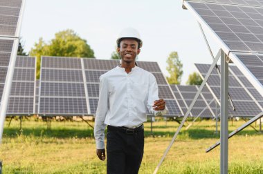 Siyah şapkalı Afrikalı mühendis güneş paneli çiftliğinde yürüyor ve gülümsüyor, yeşil enerji ve sürdürülebilirliği destekliyor.