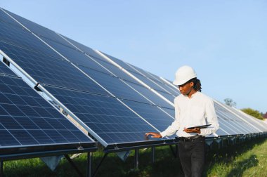 Afrikalı Amerikalı adam, mühendis, güneş panellerini inceliyor, bir güneş enerjisi çiftliğindeki dijital tablet üzerindeki verileri analiz ediyor.