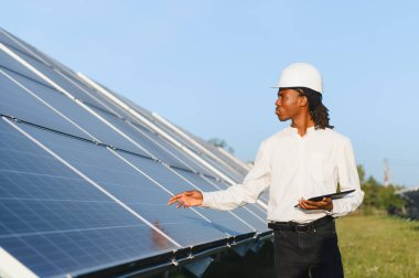 Sert şapkalı genç siyah mühendis güneş panellerini açık güneş çiftliğinde güneşli bir günde tabletle inceliyor, sürdürülebilir enerji işi.