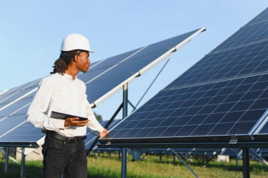 Siyah mühendis elinde tablet, yenilenebilir enerji santralindeki güneş panellerini inceliyor.