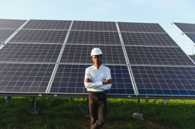 Şapkalı genç bir adam, kollarını kavuşturmuş, güneş panellerinin önünde duruyor, yenilenebilir enerjiyi temsil ediyor.