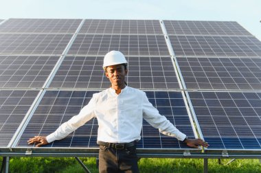 Sert şapkalı Afrikalı mühendis yenilenebilir enerji ve sürdürülebilirliği temsil eden güneş panellerinin önünde güvenle duruyor