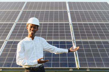 Sert şapkalı gülümseyen genç siyah mühendis açık hava güneş paneli dizisini sunuyor, temiz enerji geleceğine odaklanıyor.