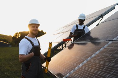 Büyük güneş panelleri takan sert şapkalı profesyonel işçiler. Altın saat boyunca yenilenebilir enerji istasyonu üzerinde takım çalışması