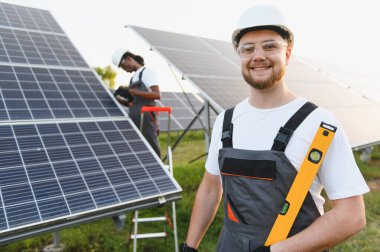 Seviyeyi korurken gülümseyen genç bir adam, arka planda çalışan bir meslektaşla güneş panellerinin önünde duruyor. Temiz elektrik üretiliyor.