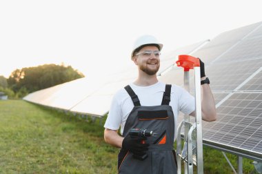 Güneş panellerinin yanında durup, yenilenebilir enerjiyi destekleyen güvenlik teçhizatı içinde gülümseyen bir işçi matkap tutuyor.