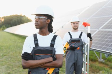 Yenilenebilir enerji işçileri güneş enerjisi santralinde sabit şapka ve güvenlik gözlüğü takarak fotovoltaik paneller kuruyorlar.