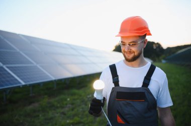 Elinde parlak bir ampul tutarken gülümseyen kasklı ve güvenlik gözlüklü teknisyen, sürdürülebilir enerji yeniliğini temsil ediyor.