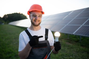 Sert şapkalı gülümseyen adam, aydınlatılmış bir ampulü işaret ederek güneş enerjisinin temiz enerjisini sembolize ediyor.