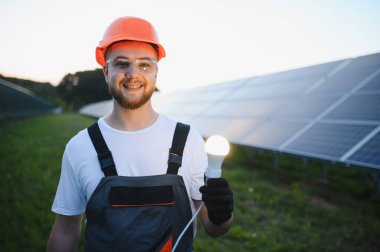 Başlık takan, güvenlik gözlüklü, gülümseyen, güneş panellerinin önünde parlayan bir ampul tutan mühendis.