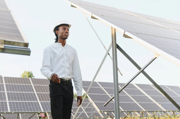 Sert bir şapka takan Afrikalı mühendis büyük bir yenilenebilir enerji alanındaki güneş panellerini geziyor ve denetliyor.