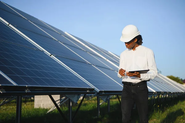Sert şapkalı adam büyük güneş paneli dizisini denetliyor, yenilenebilir enerji izleme tabletine veri kaydediyor.