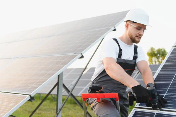 Sert bir şapka ve güvenlik gözlüğü takan erkek işçi dışarıda matkapla bir güneş paneli kuruyor ve yeşil enerji üretiyor.