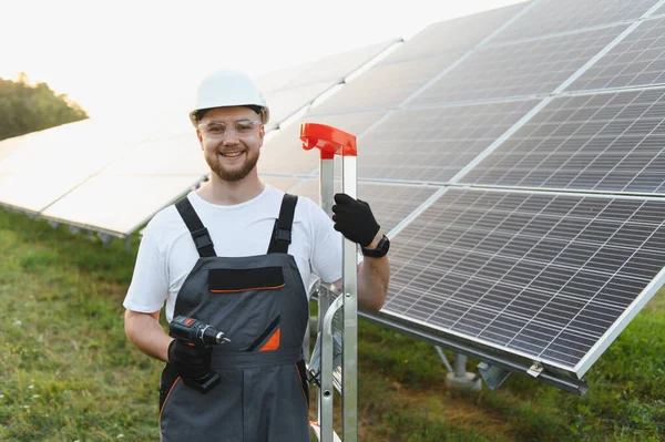 Sert şapkalı ve üniformalı gülümseyen erkek teknisyen fotovoltaik panelleri dışarı yerleştirirken merdivende matkap kullanıyor.