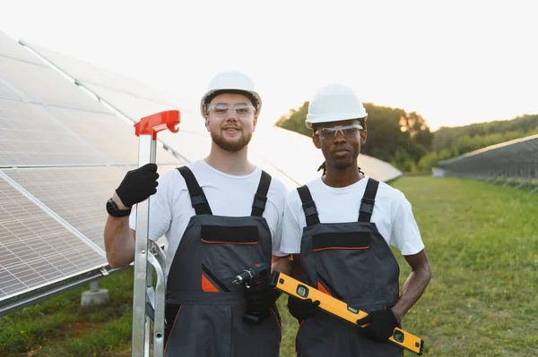 Kurulum sırasında güvenlik ekipmanı giyen iki farklı mühendis güneş paneli alanının önünde duruyor ve ellerinde aletler var.