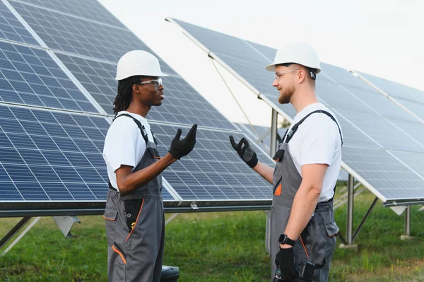 İki farklı erkek mühendis, güneş enerjisi santralinde çalışırken başlık ve güvenlik gözlüğü takıp tartışıyorlar.