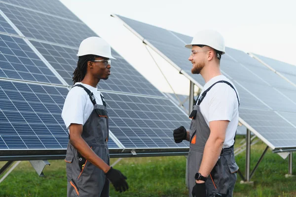 Güneş paneli alanında yenilenebilir enerji projesini tartışan iki farklı başlık ve tulumlu mühendis.